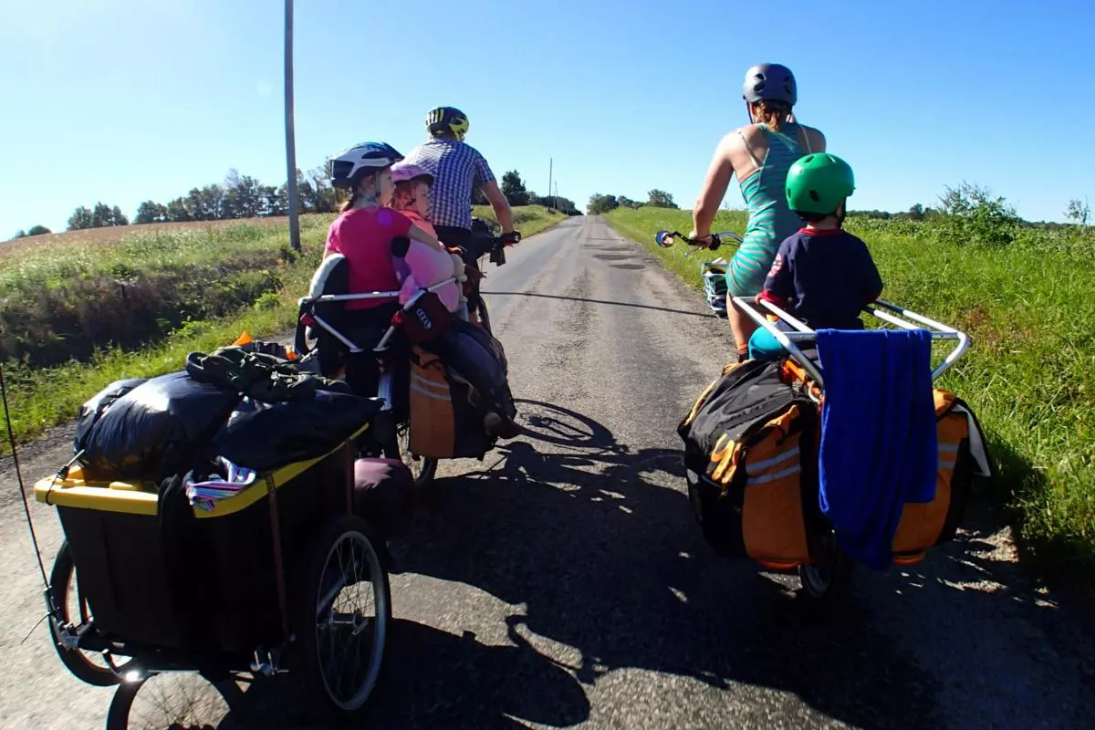 Family cycling on a rural road with two adults and two children, wearing helmets, towing loaded bike trailers.